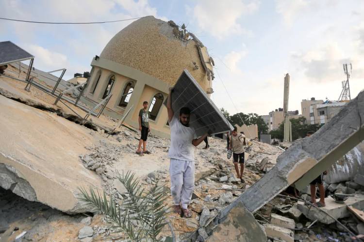 Warga Palestina mengangkat solar panel dengan latar bangunan masjid yang hancur akibat serangan Israel. (Foto: Aljazera)