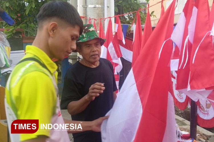Pak Nano (bertopi) begitu gembira ketika mulai pertama ada yang membeli benderanya. Ia mengaku masih optimis jualan musiman bisa untuk biaya hidup keluarga, Selasa (22/7/2025). (Foto: Sutrisno/TIMES Indonesia)